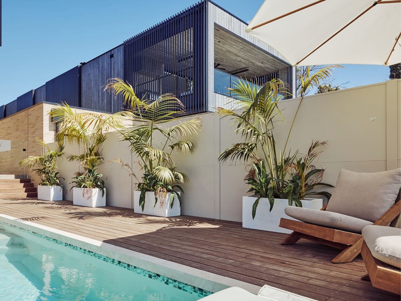House 2 takes a softer approach to poolside living. Warm timber decking, palms and pale furniture create an easy coastal feel, with sage striped cushions adding a gentle hint of colour.